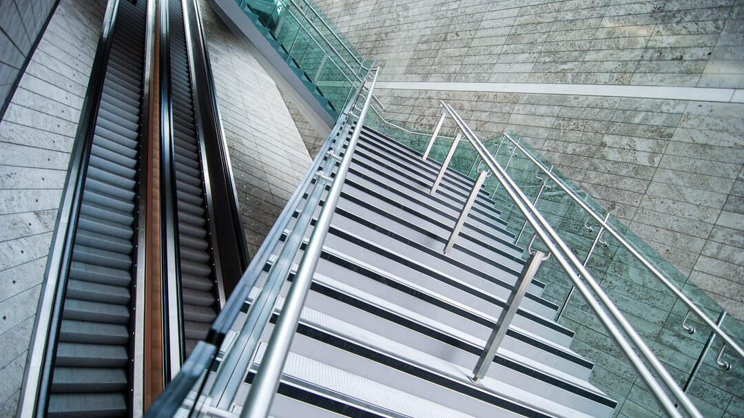 Das Foto zeigt zwei Rolltreppen sowie eine Treppe, die über die beiden Rolltreppen führt. Die Rolltreppen laufen gerade aus.  Die moderne aus Metall bestehende Treppe überquert die Rolltreppen, jedoch versetzt im Zickzack. 