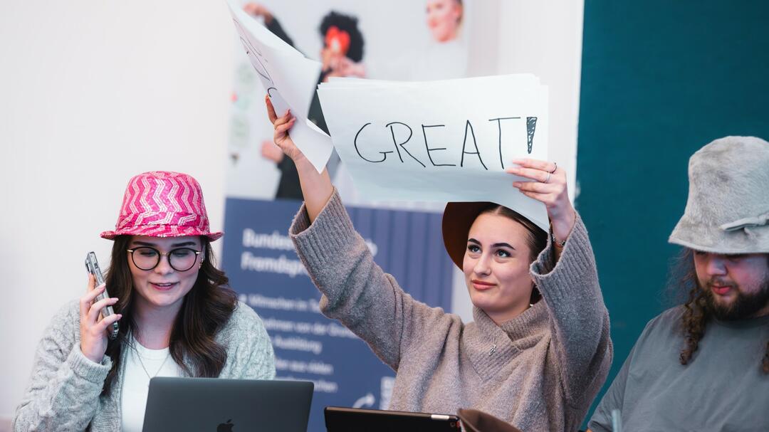 3 Teilnehmer des Azubiturniers des Bundeswettbewerb Fremdsprachen. Die junge Frau in der Mitte hält ein Schild mit der Aufschrift "Great" hoch.