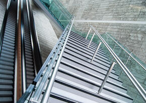 Das Foto zeigt zwei Rolltreppen sowie eine Treppe, die über die beiden Rolltreppen führt. Die Rolltreppen laufen gerade aus.  Die moderne aus Metall bestehende Treppe überquert die Rolltreppen, jedoch versetzt im Zickzack. 