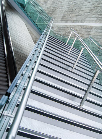 Das Foto zeigt zwei Rolltreppen sowie eine Treppe, die über die beiden Rolltreppen führt. Die Rolltreppen laufen gerade aus.  Die moderne aus Metall bestehende Treppe überquert die Rolltreppen, jedoch versetzt im Zickzack. 
