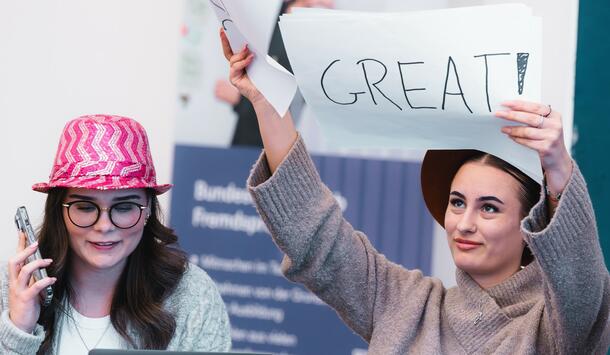 2 junge Frauen, die am Azubiturnier des Bundeswettbewerb Fremdsprachen teilnehmen. Die rechte Person hält ein Schild mit der Aufschrift "Great" hoch.